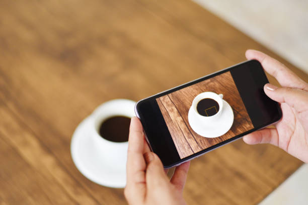 istock Taking photo coffee cup with camera smartphone for use technology post upload file to online on Internet - Close up of woman hand holding mobile phone photographing coffee cup on wooden table 1164038850