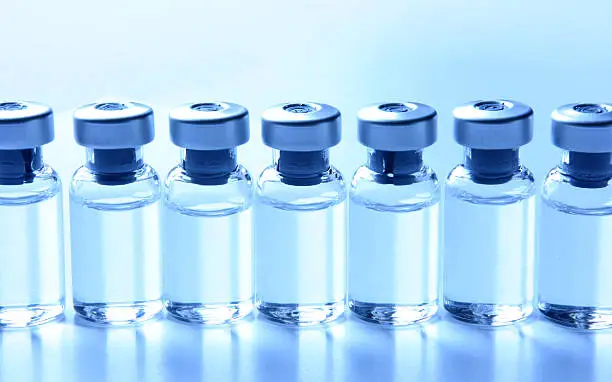 Close-up of a series of many small generic medical vials standing in a row filled with liquid medication. No labels on the seven bottles. Concepts like vaccination, flu shot. Image is blue tinted. Studio shot.