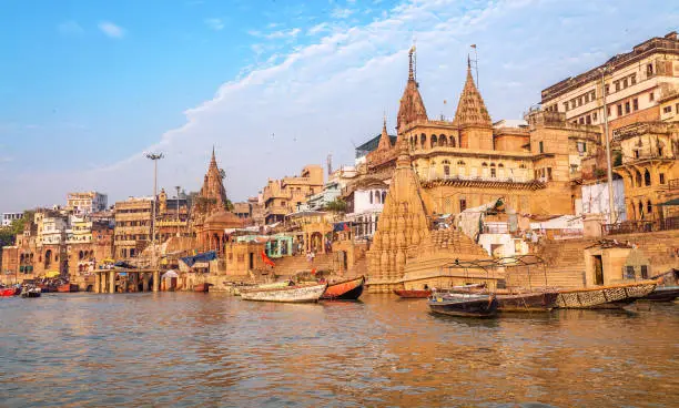 Historic Varanasi city architecture with ancient buildings and temple along the Ganges river ghat with view of wooden boat used for pleasure trips for tourists on the Ganges