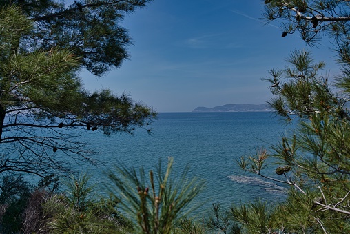 Thasos, Greece - April 29, 2019: A peninsula surrounded by The Aegean Sea water seen through a green opening eye of trees with the shore view in the distance, in Thasos, Greece