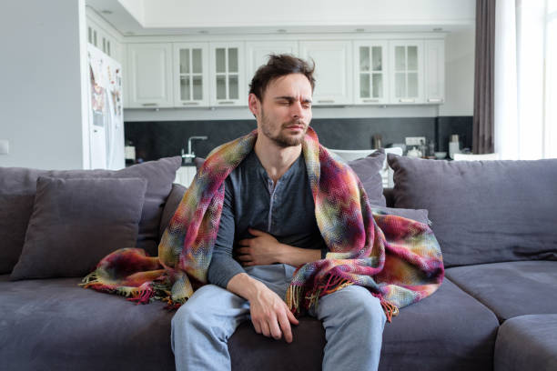 Ailing man A sick man sits on a sofa in blankets and holds his stomach cold and flu man stock pictures, royalty-free photos & images