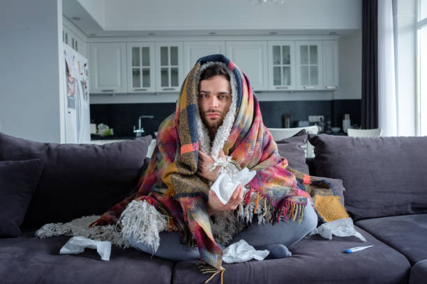 The man with the temperature at home A man with a temperature in plaids sits on the couch, a thermometer in his mouth cold and flu man stock pictures, royalty-free photos & images