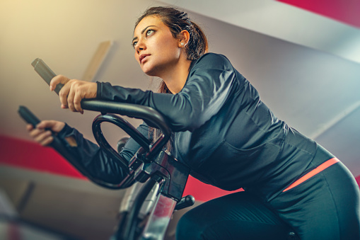Indoor image of an Asian/Indian, beautiful mid adult woman does cardio exercise on an air bike in a gymnasium.