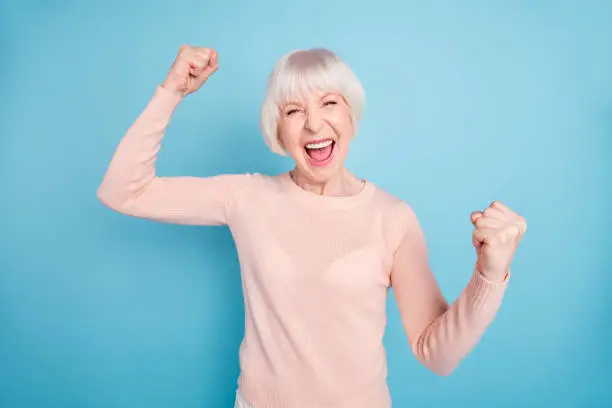 Photo of Portrait of cheerful lady shouting yeah raising fists wearing pastel sweater isolated over blue background