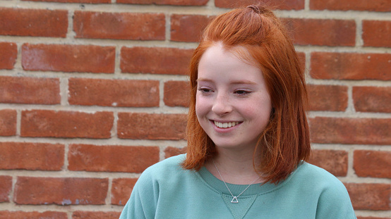 Stock photo of young redhead girl 13 / 14 years old looking sideways, red brick wall background. This teenage girl has short bob hairstyle and red ginger hair tied back, smiling perfect teeth after dentist wearing brace with green sweatshirt, silver triangle pendant necklace
