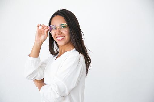 Cheerful beautiful woman looking at camera through trendy eyeglasses. Young Latin woman in white shirt adjusting eyeglasses. Visionary concept