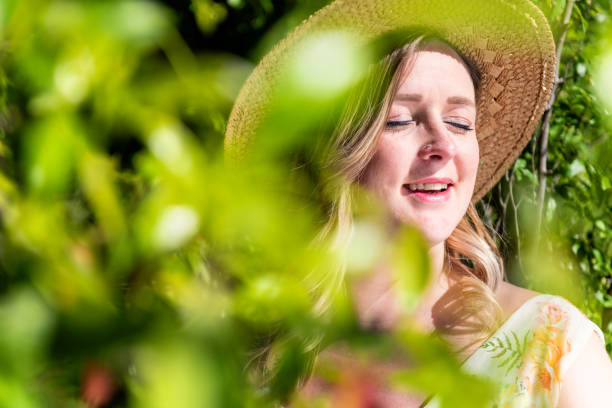 Pretty woman in nature relaxing with eyes closed and smiling. Young woman standing behind branch, wearing straw hat and relaxing. Focus on woman, branch of plant defocused.