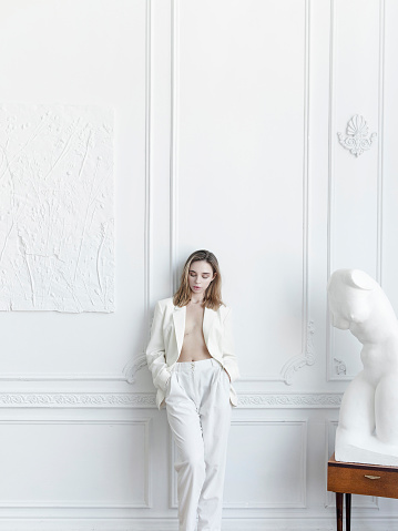 Luxury lifestyle Beautiful young woman wearing white suit: pants and blazer hands in pockets She is Parisian apartment Looks gorgeous confident with fresh natural make-up Amazing vintage style interiour White walls sculpture unique furniture
