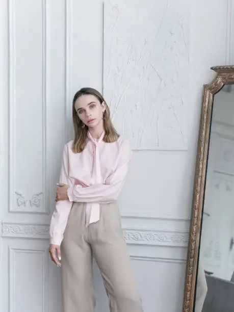 Luxury lifestyle Beautiful young woman wearing vintage clothing pink blouse beige wide leg pants She is Parisian apartment standing next to antique mirror Looks gorgeous confident with fresh natural make-up Amazing interiour  Sophisticated life