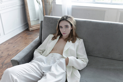Luxury lifestyle Beautiful young woman wearing white suit: pants and blazer in Parisian apartment Looks gorgeous Fresh natural make-up Fashion model lying or sitting on the gray color sofa Amazing vintage style interiour