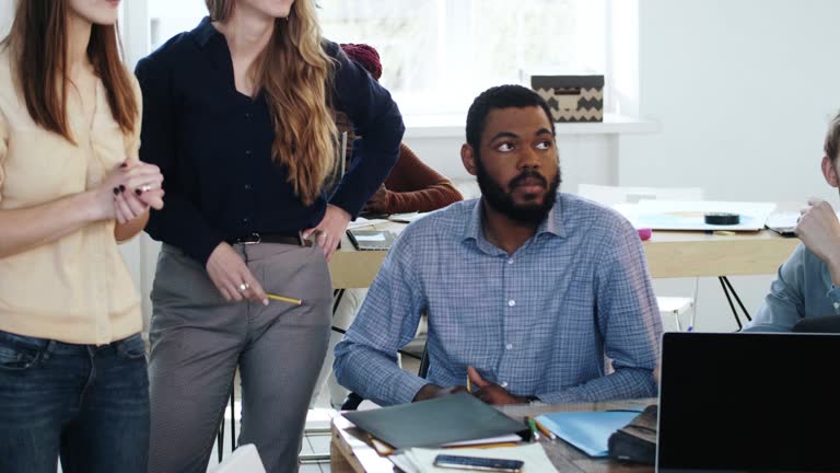 Stressed and upset African male employee sitting at modern office table busy with paperwork near two female colleagues.