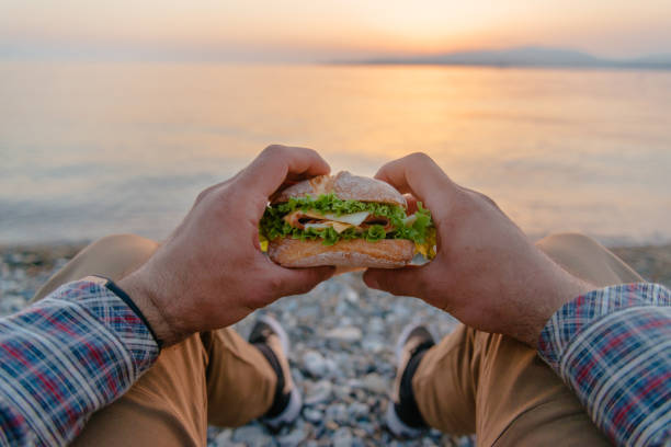 Young man resting with burger by the sea, pov. stock photo