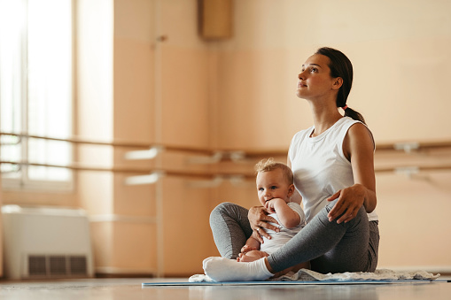 Young mother sitting in lotus position while meditating with her baby in health club. Copy space.