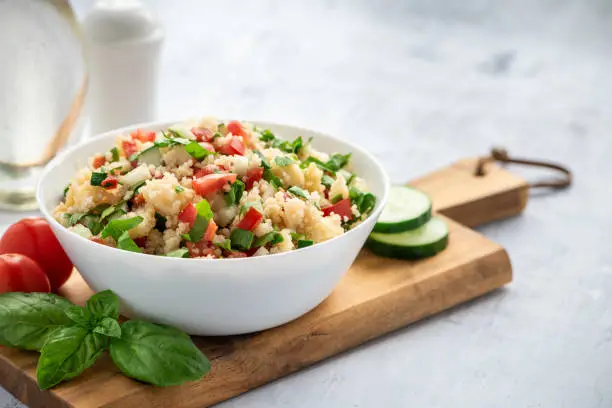 Tabbouleh - Arabian vegetarian salad made of couscous, tomatoes, cucumbers, parsley, onions and lemon juice. Couscous with vegetables in a white bowl on the table.