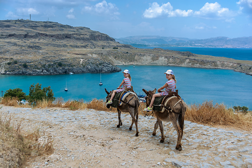 Two twin girls riding donkeys on a mountain road to the sea. Lindos. the island of Rhodes. Greece. Sunny summer day