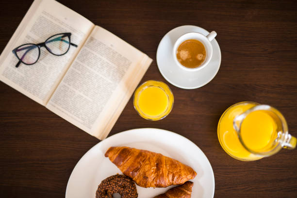 Beautiful Caucasian woman wearing eyeglasses and domestic dress having breakfast at home and reading book.