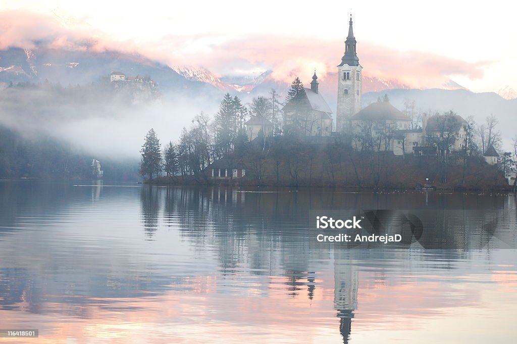 Misty Evening Reflection The Bled island church reflecting in the water with the snowy mountains and the Bled castle in the background Lake Bled Stock Photo