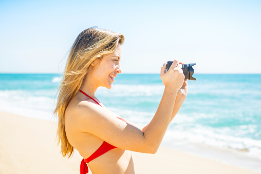 Happy young woman in red bikini taking photos on the beach in her holidays.