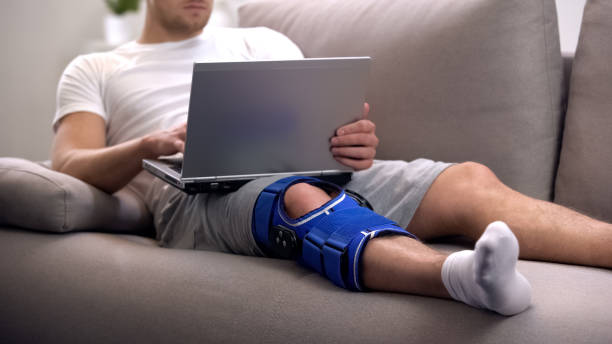 Man in arthritis knee brace working on laptop at home rehab period and freelance Man in arthritis knee brace working on laptop at home rehab period and freelance neoprene photos stock pictures, royalty-free photos & images