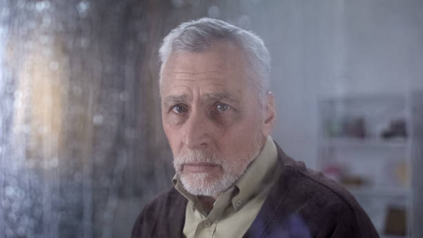 istock Lonely sad grandfather looking in camera through rainy window, nursing home 1164204636