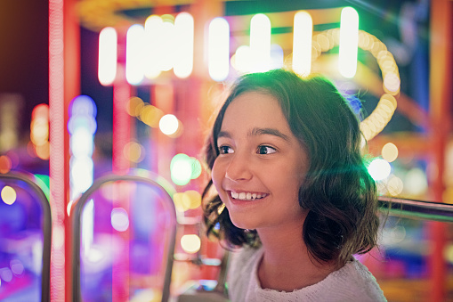 Adorable little girl near the carousel outdoors in park. Happy kid in a hat standing with hands to the side. Family weekend or vacations. Summer concept