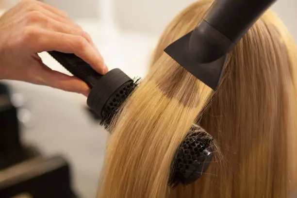 Cropped close up of a professional hairdresser using round brush and blow dryer, styling long blond hair of his female client. Healthy strong hair of a woman during new hairstyle treatment, copy space