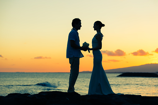 Romantic senior couple silhouettes hugging and looking at each other at coastline at sunset, side view medium shot
