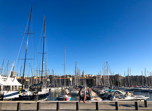 Marseille, France: Two young women walking in early morning at Marseille’s sunny Old Port/Vieux Port, with sailboats and cityscape n the background. Copy space in the vibrant blue sky.