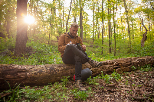 one young man, in forest, holding a flower in his hands, writing down his notes on it.