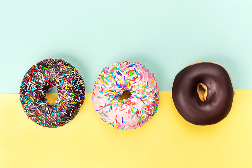 Creative shot of three tasty doughnut with colorful sprinkles on turquoise and yellow background. Flat lay, top view.