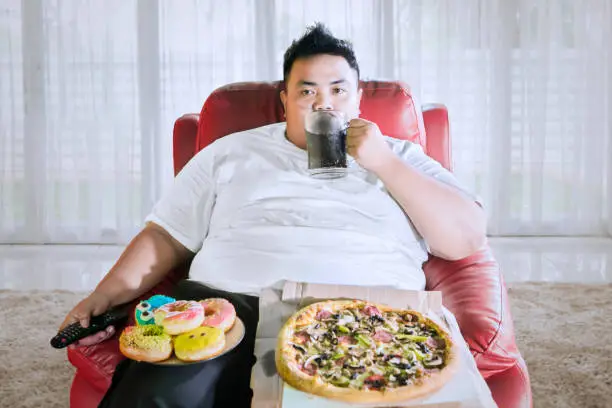 Fat man drinking a glass of cola and eating junk foods while watching television on the sofa. Shot at home