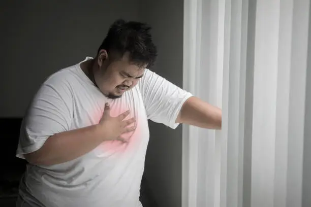 Image of overweight man having a heart attack while standing near the window in the dark room