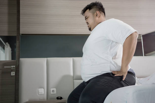 Overweight man having backache on the bed Overweight man having backache while sitting on the bed. Shot at home coccyx photos stock pictures, royalty-free photos & images