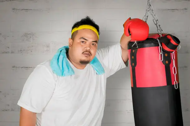 Picture of an overweight man looking at the camera while leaning on a boxing bag with brick wall background
