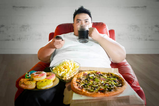 joven gordo disfrutando de comida chatarra en el sofá - comer demasiado fotografías e imágenes de stock