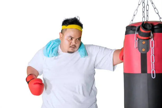 Image of overweight man hitting a boxing bag and exercising in the studio, isolated on white background
