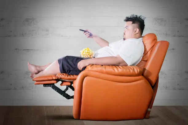 Side view of obese man eating popcorn on the sofa while watching television at home