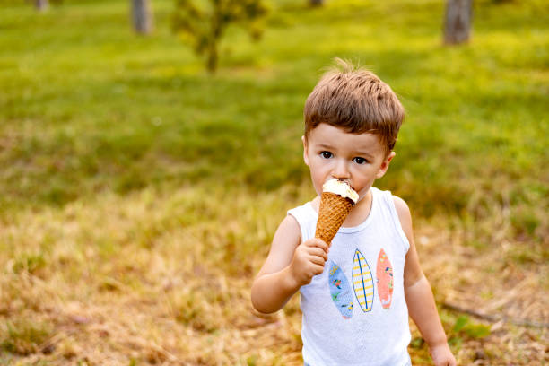 Cute little boy eating ice cream in the park Little toddler baby boy eating ice cream in cone in summer. Portrait of Adorable cute little brown hair Caucasian boy eating ice cream bar in a park, messed up and dirty, hot sunny summer day. Messy Boy standing and eats chocolate ice cream while looking at the camera. Beautiful little boy eating ice cream in cone in summer. licking ice cream cone stock pictures, royalty-free photos & images