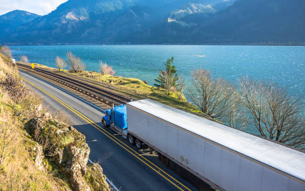 Big rig blue semi truck with refrigerated semi trailer transporting goods running on the road along railroad in Columbia Gorge Big rig classic American powerful blue semi truck with refrigerated semi trailer transporting frozen goods running on the road along railroad and river in Columbia Gorge area with mountain ranges car transporter stock pictures, royalty-free photos & images