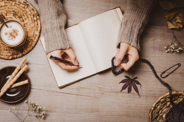 istock Fall winter background. Female hand holding a pen for a sketch or letter on old notebook with a ball of wool yarn and cup of cappuccino on a wooden background. Home hobbies, Cozy comfort concept. 1164268660