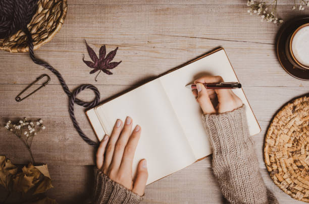 istock Fall winter background. Female hand holding a pen for a sketch or letter on old notebook with a ball of wool yarn and cup of cappuccino on a wooden background. Home hobbies, Cozy comfort concept. 1164268691