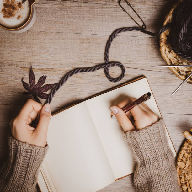 istock Fall winter background. Female hand holding a pen for a sketch or letter on old notebook with a ball of wool yarn and cup of cappuccino on a wooden background. Home hobbies, Cozy comfort concept. 1164268719