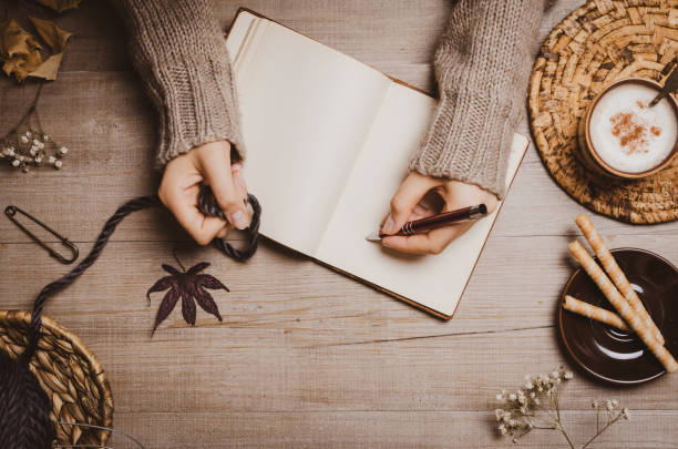 istock Fall winter background. Female hand holding a pen for a sketch or letter on old notebook with a ball of wool yarn and cup of cappuccino on a wooden background. Home hobbies, Cozy comfort concept. 1164268734