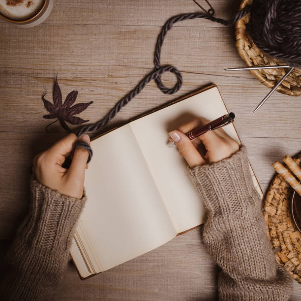 istock Fall winter background. Female hand holding a pen for a sketch or letter on old notebook with a ball of wool yarn and cup of cappuccino on a wooden background. Home hobbies, Cozy comfort concept. 1164268759
