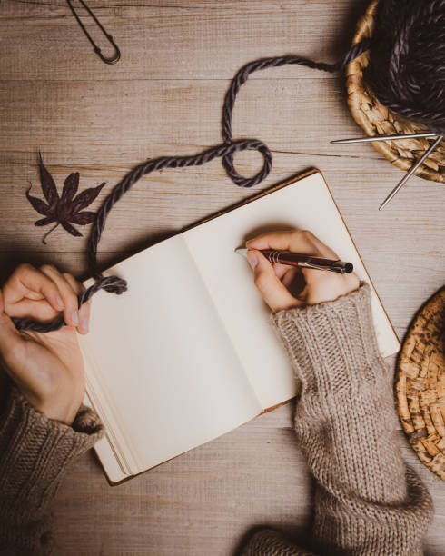 istock Fall winter background. Female hand holding a pen for a sketch or letter on old notebook with a ball of wool yarn and cup of cappuccino on a wooden background. Home hobbies, Cozy comfort concept. 1164268772