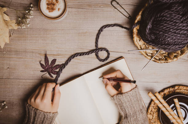 Fall winter background. Female hand holding a pen for a sketch or letter on old notebook with a ball of wool yarn and cup of cappuccino on a wooden background. Home hobbies, Cozy comfort concept. fondo de invierno de otoño. mano femenina sosteniendo un bolígrafo para un boceto o una letra en un cuaderno viejo con una bola de hilo de lana y una taza de capuchino sobre un fondo de madera. pasatiempos caseros, concepto de comodidad acogedor.
