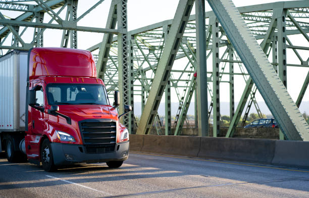 Bonnet red big rig day cab semi truck transporting cargo in dry van semi trailer driving on the truss bridge stock photo