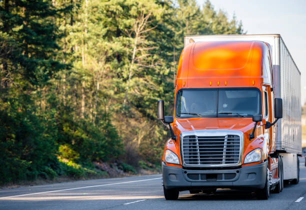 Orange big rig semi truck transporting cargo in semi trailer running on the road with trees stock photo