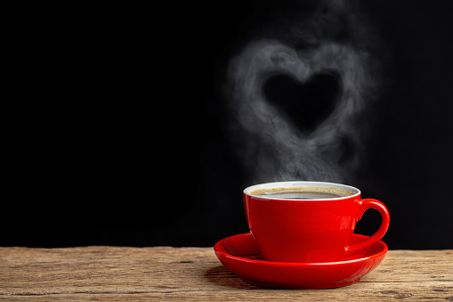 Heart of steam hovering over a red coffee cup of coffee on wooden table with black background.