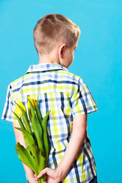 Holiday mother's day concept. Little boy has prepared surprise present for mum, flowers yellow tulips, holds it behind the back on blue